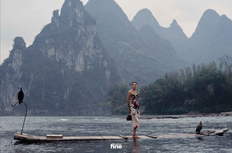 A woman in a floral dress stands on a bamboo raft on a river with mountains in the background, creating a serene and fashionable scene.