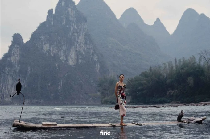 A woman in a floral dress stands on a bamboo raft on a river with mountains in the background, creating a serene and fashionable scene.