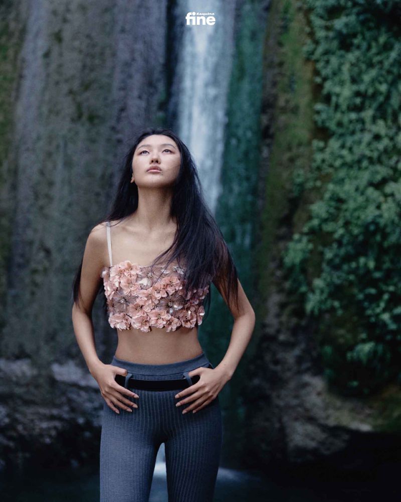 Asian woman in floral top and grey leggings poses in front of a waterfall and lush greenery, looking upward. Fashion and nature blend.