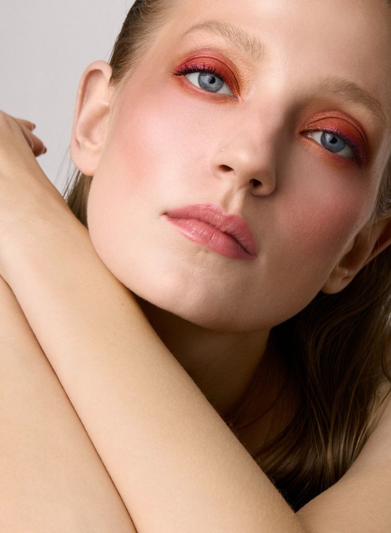Close-up beauty portrait of a woman with warm reddish-orange eyeshadow and pink lips, resting head on arms