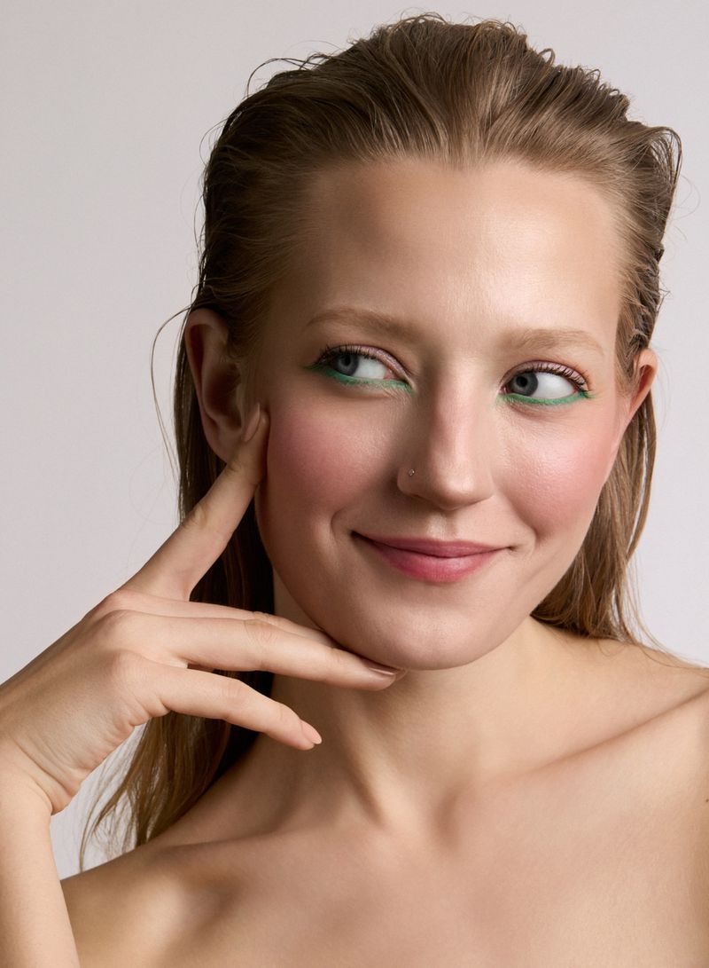 Close-up of an adult woman with light skin, light hair, pink lips, and green eye makeup smiling softly against a neutral background.