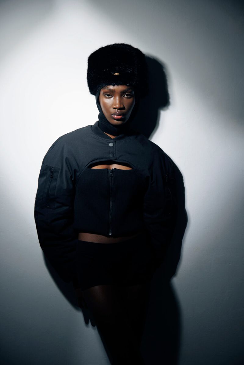 Portrait of an adult woman with dark skin wearing a black ushanka-style hat, turtleneck, and jacket against a gradient white background.