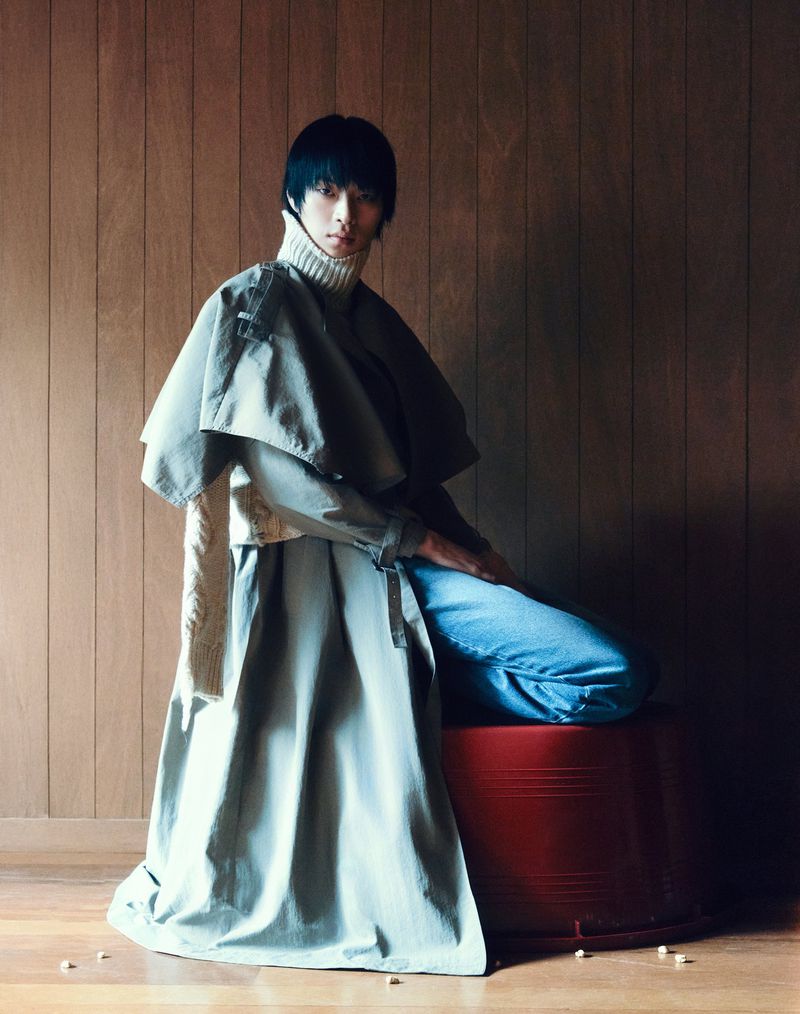Asian male in stylish overcoat poses while seated on a red stool, set against a wood panel backdrop in a fashion photography setting.