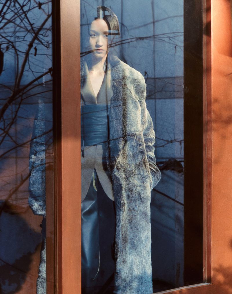 Fashion shot of an Asian adult woman wearing a furry coat behind a glass window with bare tree branches during daytime with natural lighting.