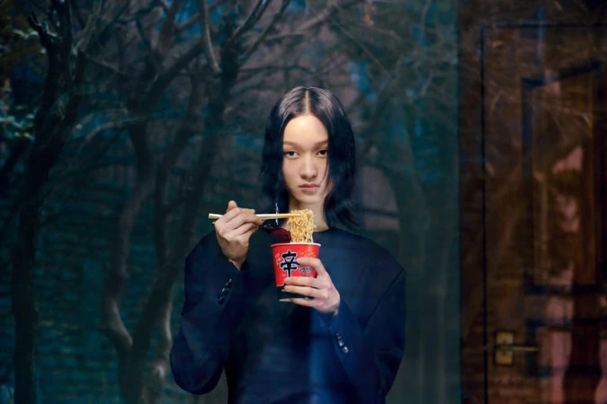 A person with long dark hair eats noodles from a cup with chopsticks, wearing a black oversized jacket in front of a blurry background.