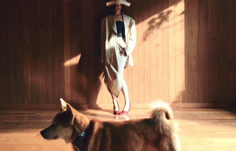 Adult female model posing in oversized tan jacket and skirt near a dog in a sunlit room with wood paneled walls and floor.