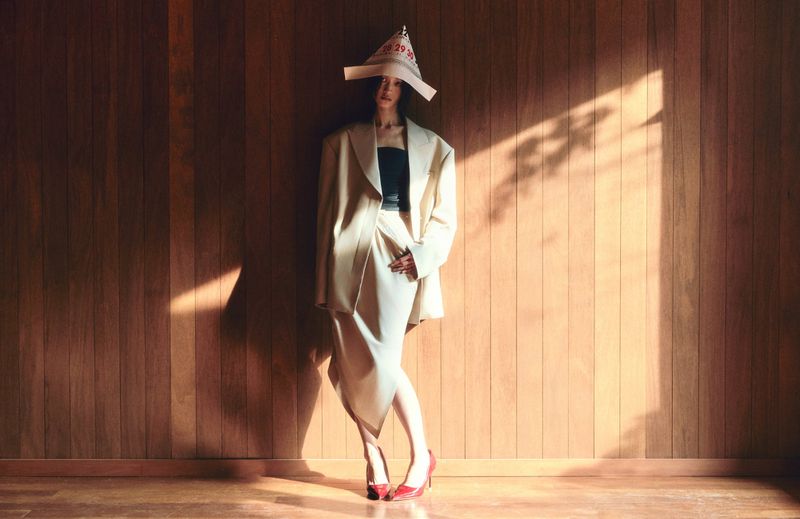 Woman posing in beige blazer, skirt, red shoes, paper hat in front of wood wall, in warm light and shadow, fashion studio shot.