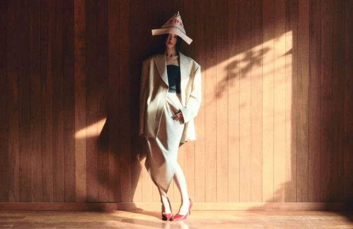 Woman posing in beige blazer, skirt, red shoes, paper hat in front of wood wall, in warm light and shadow, fashion studio shot.
