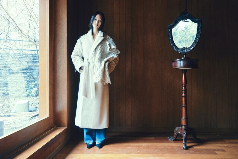 Model wearing a long white belted coat standing indoors near a large window and a vintage standing mirror.