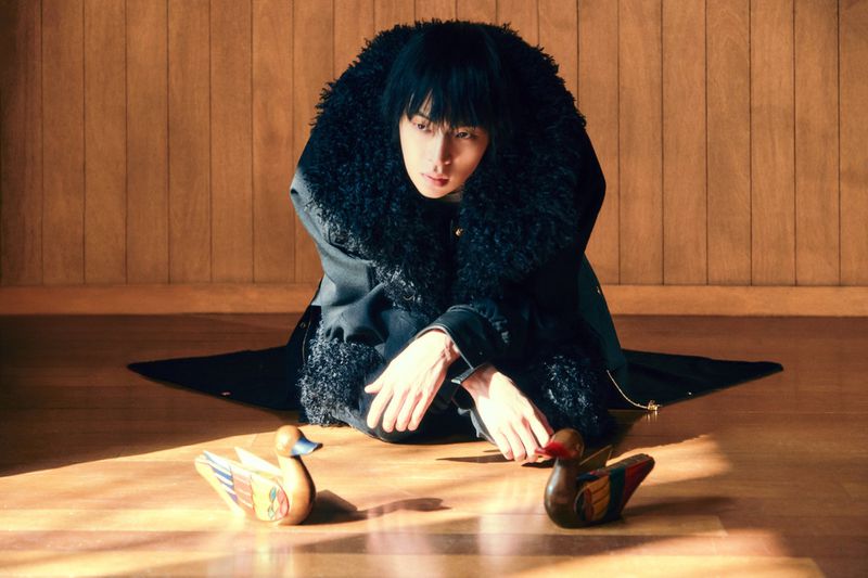 Portrait of a person with dark hair, seated on a wood floor, wearing a dark coat with fur collar, two wooden duck figures in foreground.