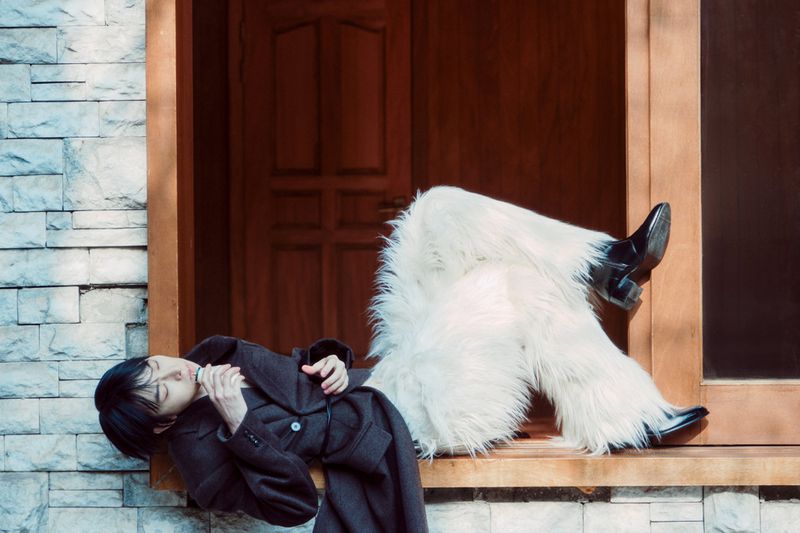 Adult lying in a window wearing a black coat and furry white pants with brick and wood wall backgrounds in an indoor environment.