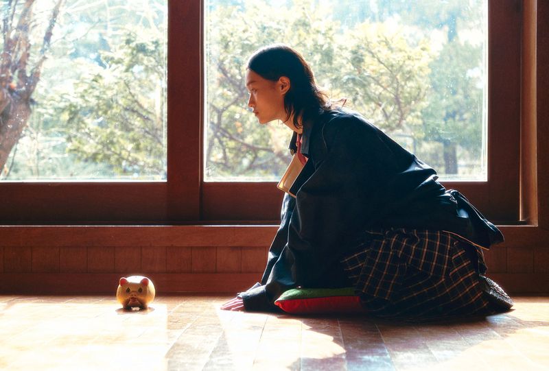 Adult male sitting indoors near a window with a piggy bank, wearing dark clothing with a plaid blanket and colored cushions.