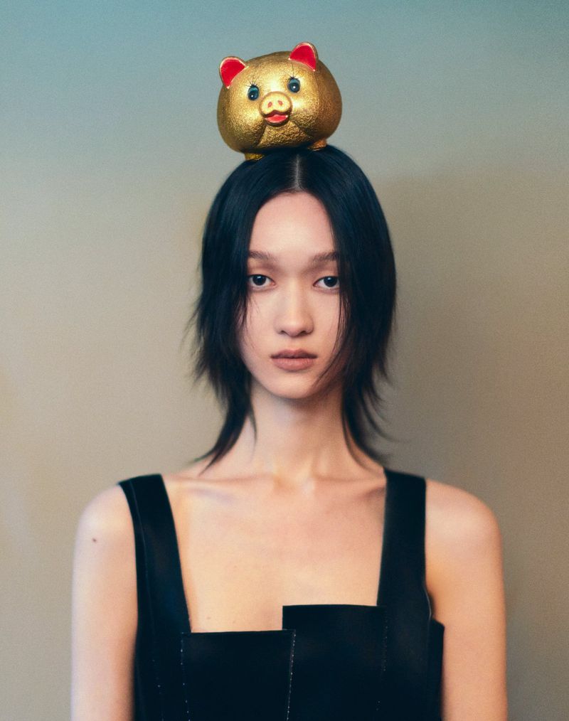 Young woman with chin-length dark hair and black top gazes at the camera with a golden pig-shaped coin bank balanced on her head.
