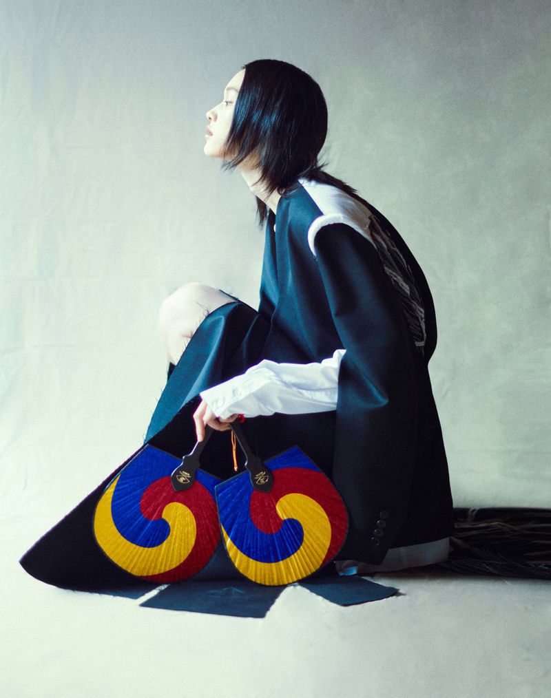 Woman in oversized blazer sitting indoors, holding a pair of colorful spiral fans. Fashion editorial photography with pale grey background.