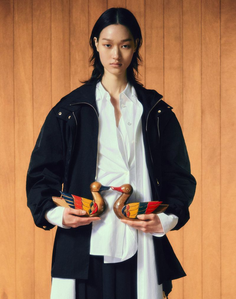 Asian woman in a black jacket and white shirt holding wooden ducks, posing in front of a wood-paneled background, fashion shot.