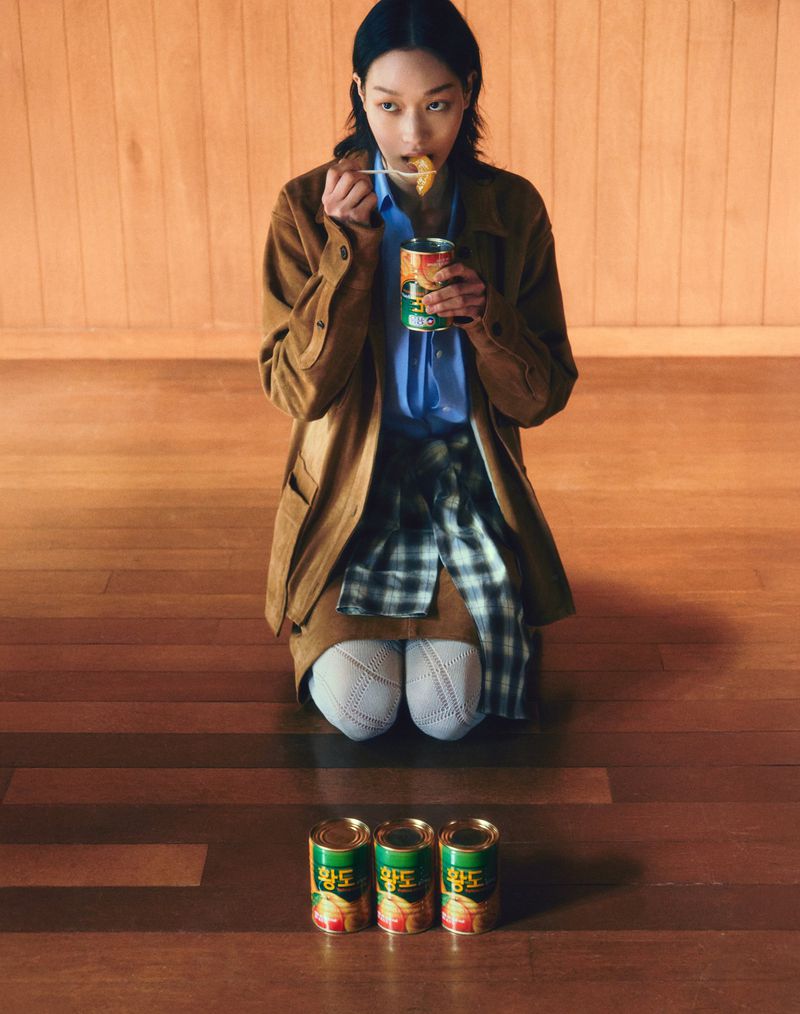 Woman in coat eats canned food on floor, three unopened cans beside her, in an indoor setting with wood floor and wall.