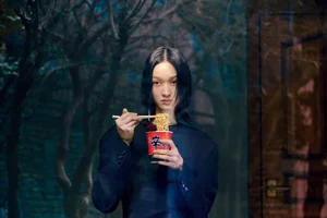 Model holding a cup of instant noodles with chopsticks in front of a dark wooded background for Marie Claire Korea editorial