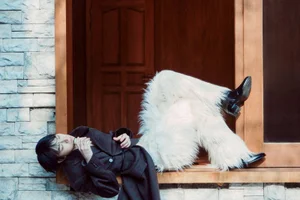 Model lying horizontally against a stone wall with white furry trousers and a dark coat