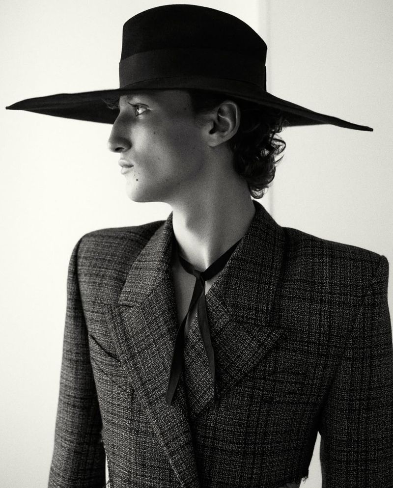 Black-and-white portrait of a male model wearing a large hat and a tweed blazer, gazing to the side in a fashion-editorial setting.