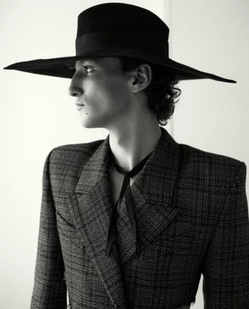 Black-and-white portrait of a male model wearing a large hat and a tweed blazer, gazing to the side in a fashion-editorial setting.