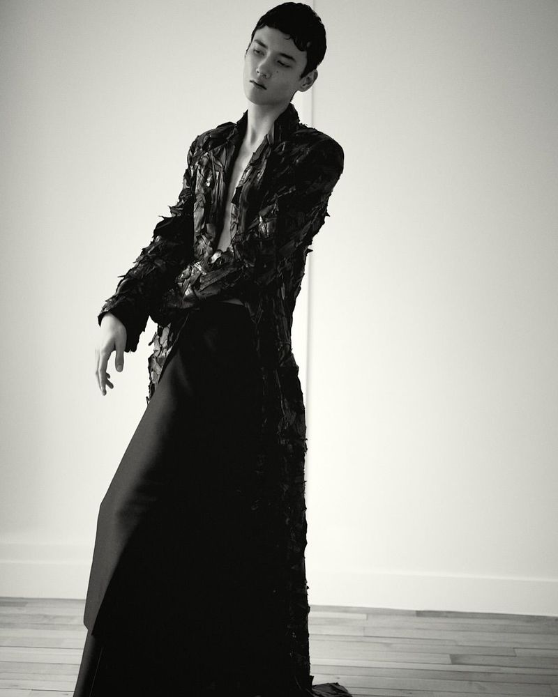 Monochrome fashion shot of an adult male in an open black textured jacket and long black skirt, posing against a white wall and wood floor.