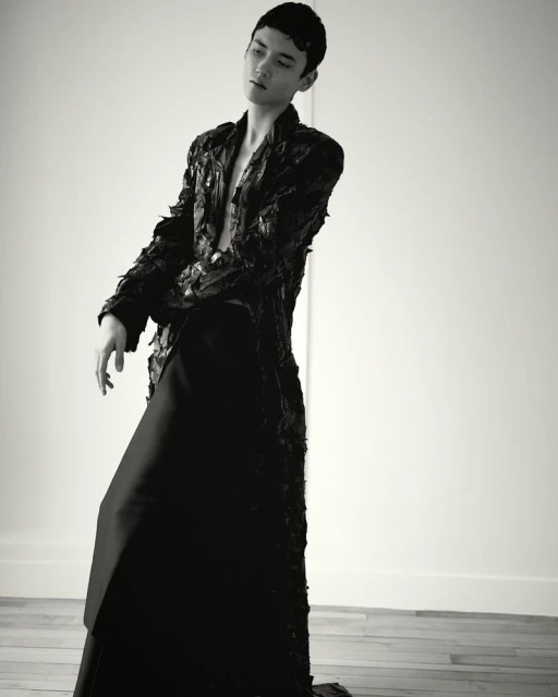Monochrome fashion shot of an adult male in an open black textured jacket and long black skirt, posing against a white wall and wood floor.