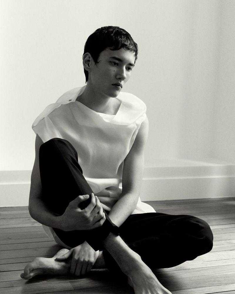 Portrait of a young adult male sitting cross-legged on a wooden floor wearing a white top and dark pants in a minimalist setting.
