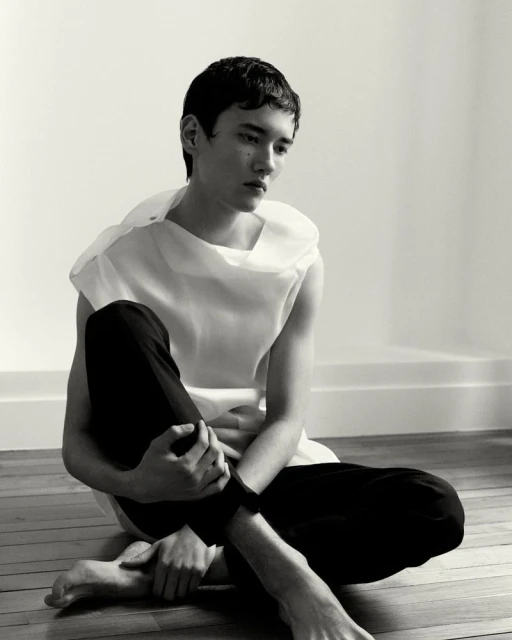 Portrait of a young adult male sitting cross-legged on a wooden floor wearing a white top and dark pants in a minimalist setting.