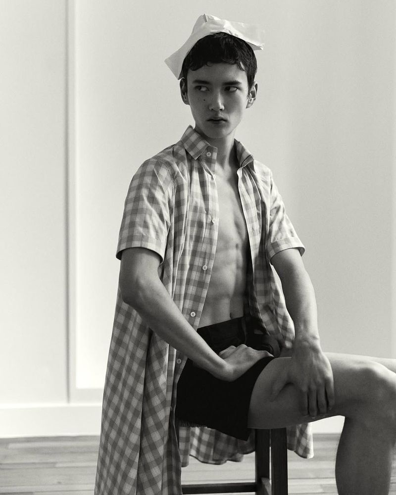 Monochrome portrait of a male fashion model in an open checkered shirt sitting on a stool with a paper accessory on his head.