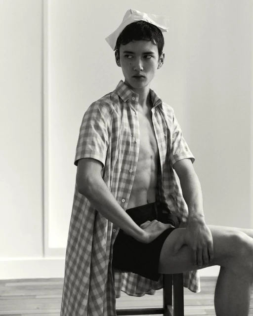 Monochrome portrait of a male fashion model in an open checkered shirt sitting on a stool with a paper accessory on his head.
