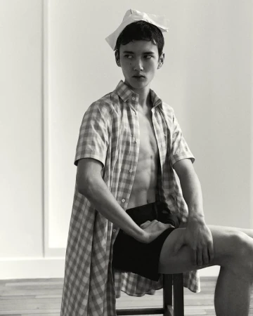 Monochrome portrait of a male fashion model in an open checkered shirt sitting on a stool with a paper accessory on his head.