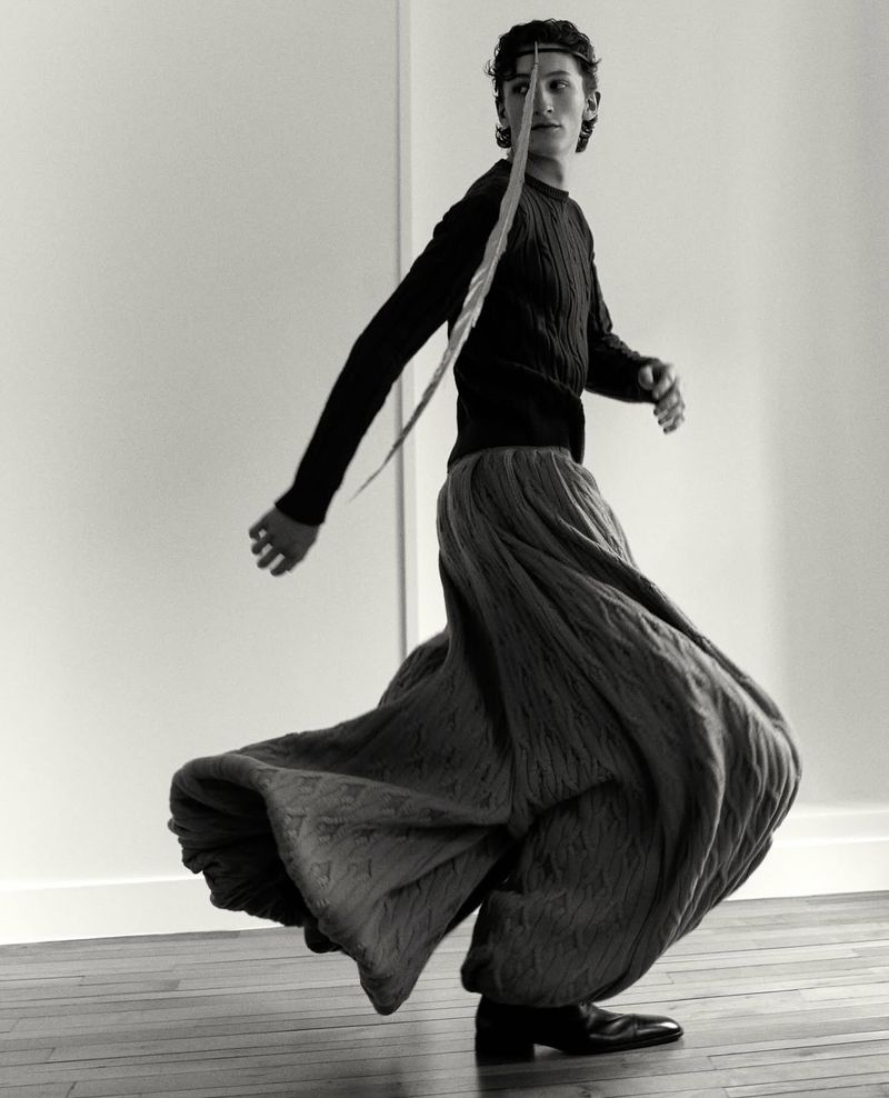 Monochrome portrait of a slender person spinning with a long feather in hair, wearing a sweater and wide skirt, indoors.