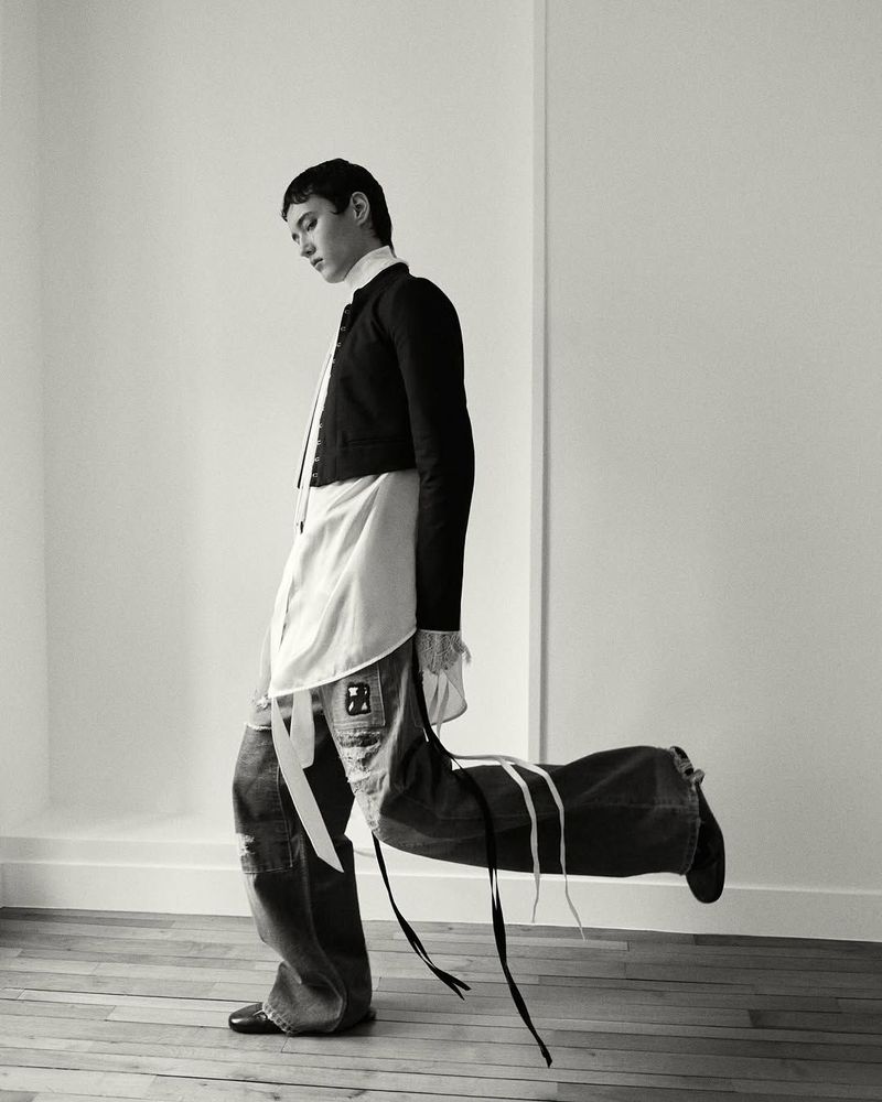 A stylishly dressed person carries a large, duffel-style bag over their shoulder indoors against a neutral wall, in a black and white fashion photo.