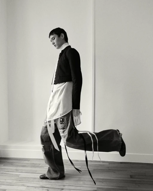 A stylishly dressed person carries a large, duffel-style bag over their shoulder indoors against a neutral wall, in a black and white fashion photo.