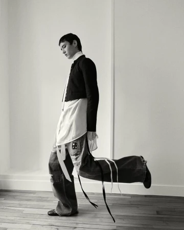 A stylishly dressed person carries a large, duffel-style bag over their shoulder indoors against a neutral wall, in a black and white fashion photo.