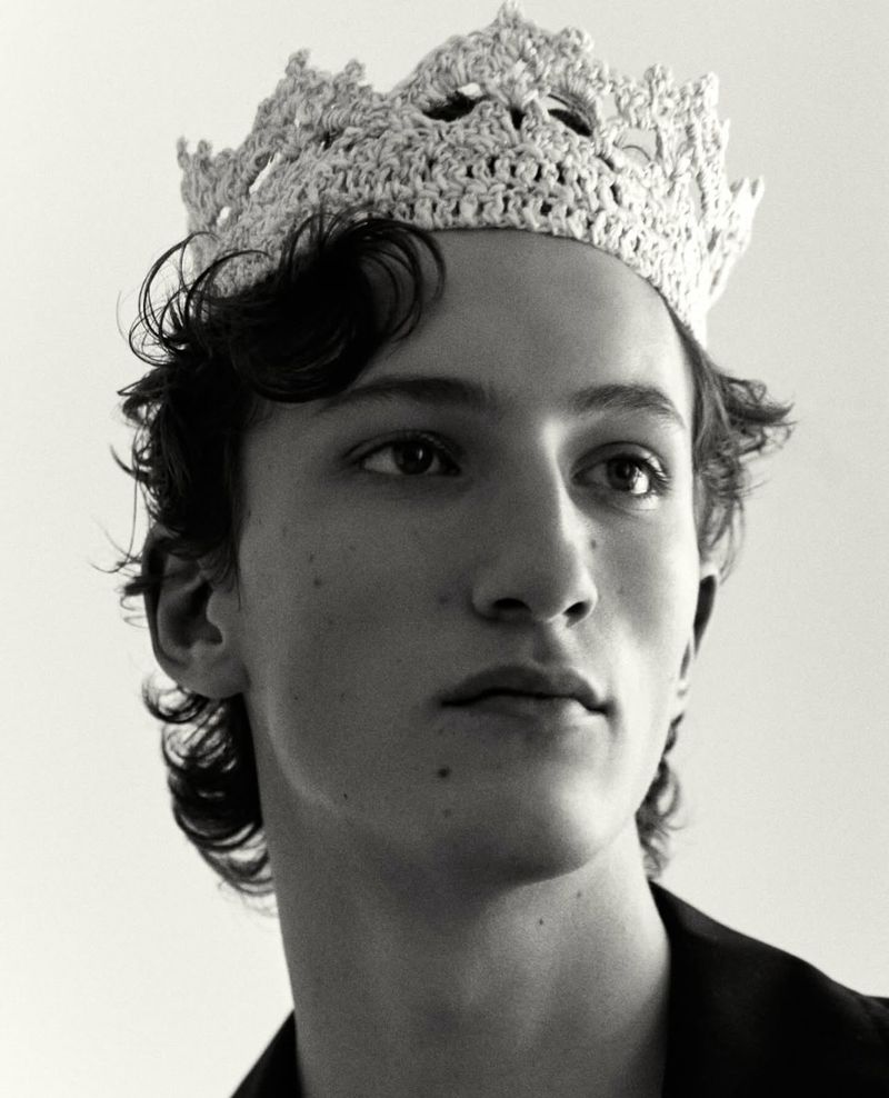 Portrait of a young adult male with dark hair wearing a knitted white crown, set against a light background in a close-up shot.