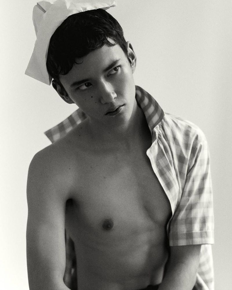 Monochrome portrait of a bare-chested young adult male looking to the side with a crumpled paper hat, wearing a plaid shirt.