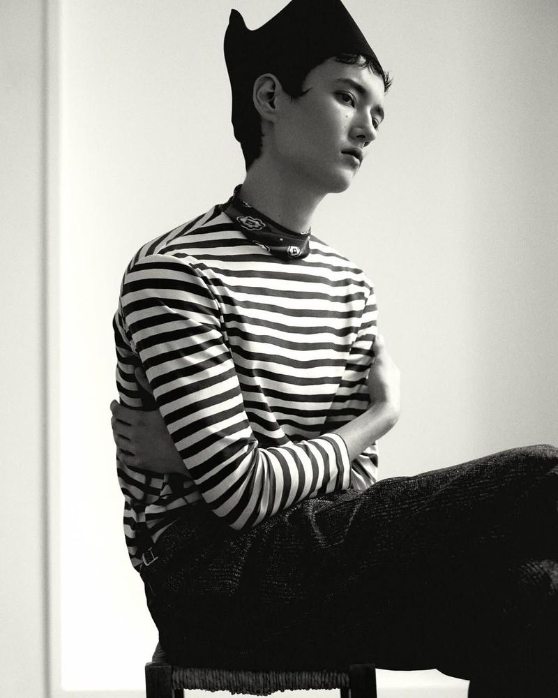 Portrait of a seated model wearing a striped long-sleeve top and a dark cap against a plain white background.