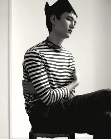 Portrait of a seated model wearing a striped long-sleeve top and a dark cap against a plain white background.