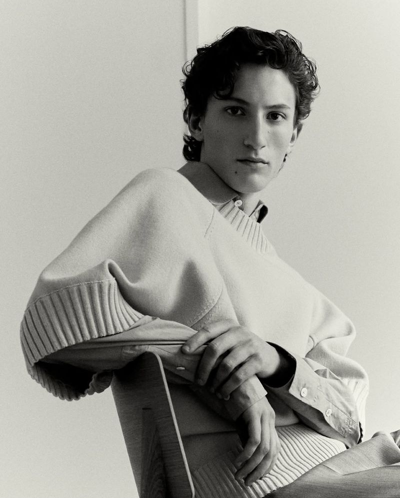 Portrait of a young male model seated backwards on a wooden chair against a plain light studio background, captured in monochrome.