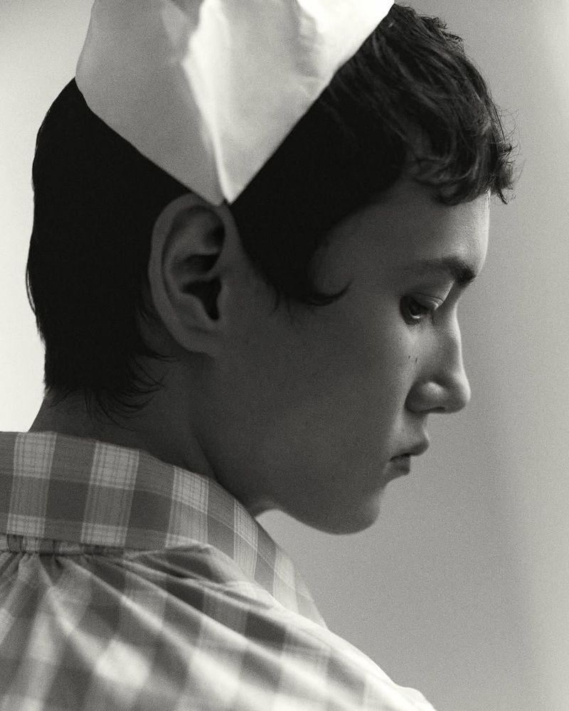 Monochrome portrait of a young adult in a checkered shirt and white cap, gazing to the side with soft studio lighting and neutral backdrop.
