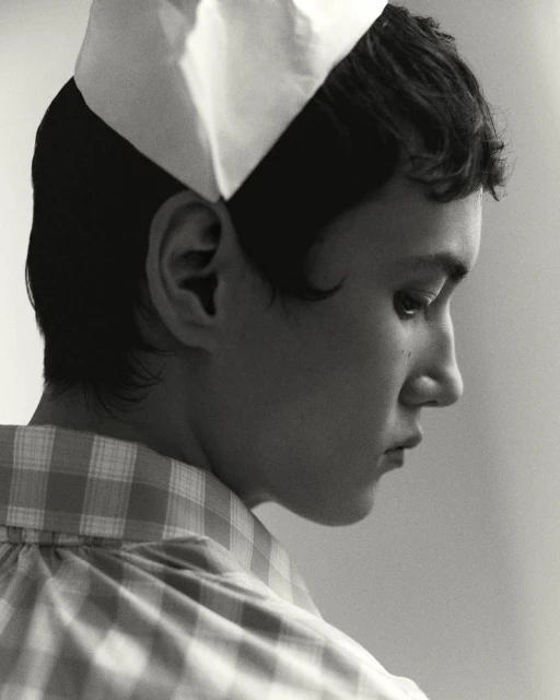 Monochrome portrait of a young adult in a checkered shirt and white cap, gazing to the side with soft studio lighting and neutral backdrop.
