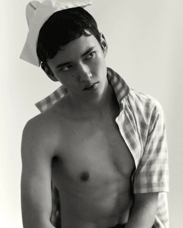 Monochrome portrait of a young male model wearing a paper hat and an unbuttoned checkered shirt against a seamless background
