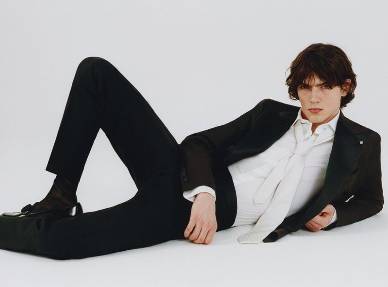 A stylish young man in a black suit and white shirt poses reclining against a white backdrop, exuding a fashionable and relaxed vibe.