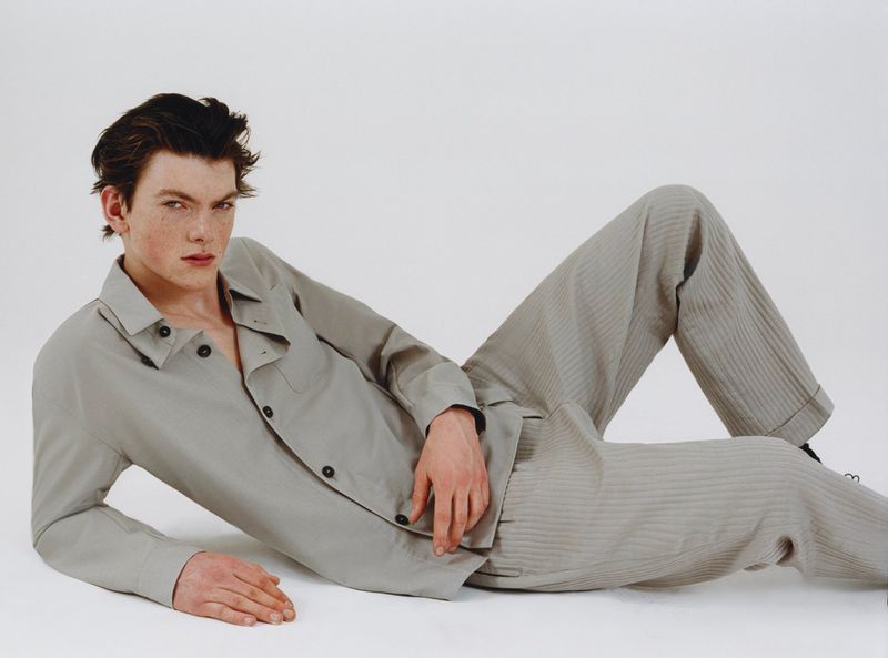 A young male model lying on a white seamless background wearing a grey button-up shirt and matching grey trousers.