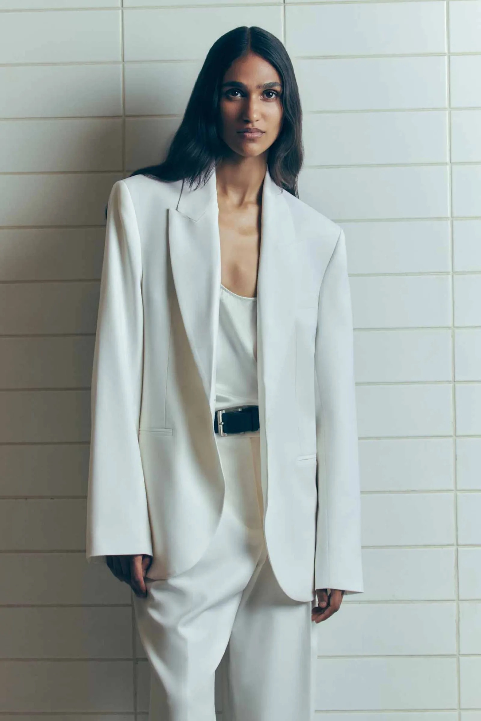 Portrait of a dark-haired woman wearing a white blazer and trousers, posing against a white tiled wall in a minimalist setting.