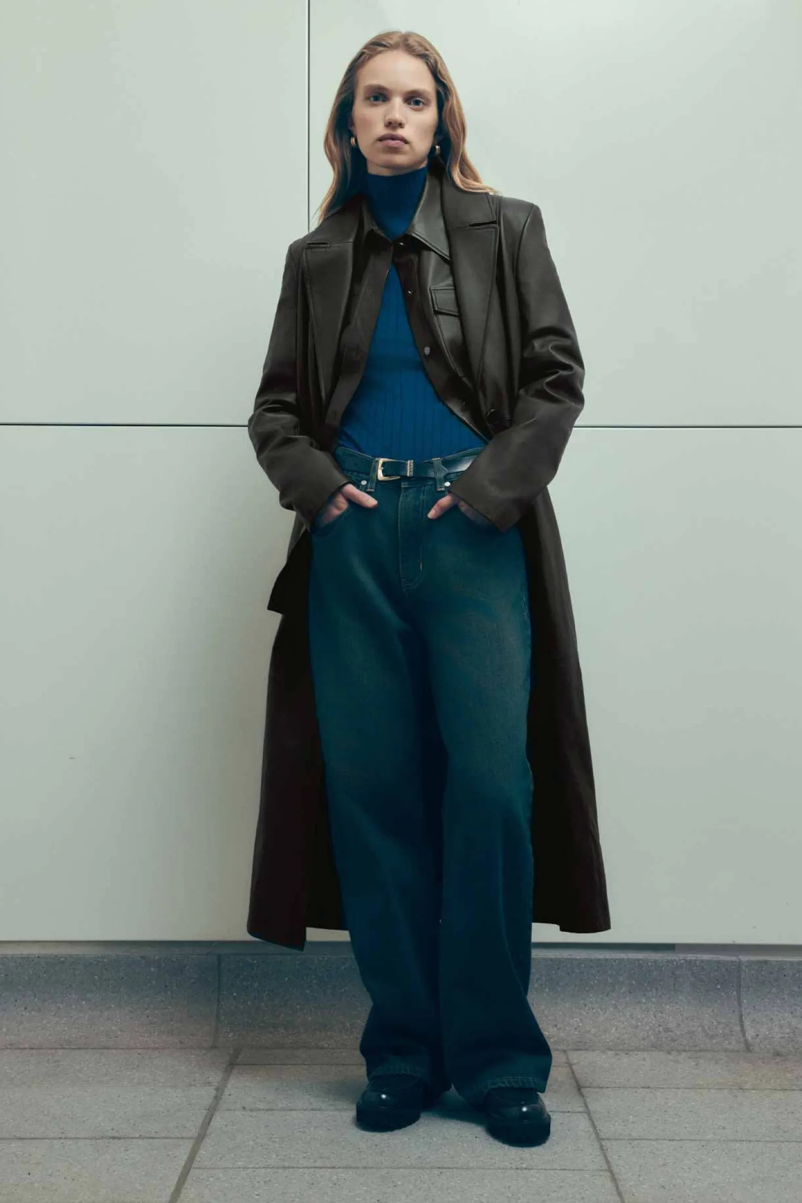 Full-length portrait of a young adult woman with long hair wearing a long brown leather trench coat, blue turtleneck, blue jeans, and black shoes.