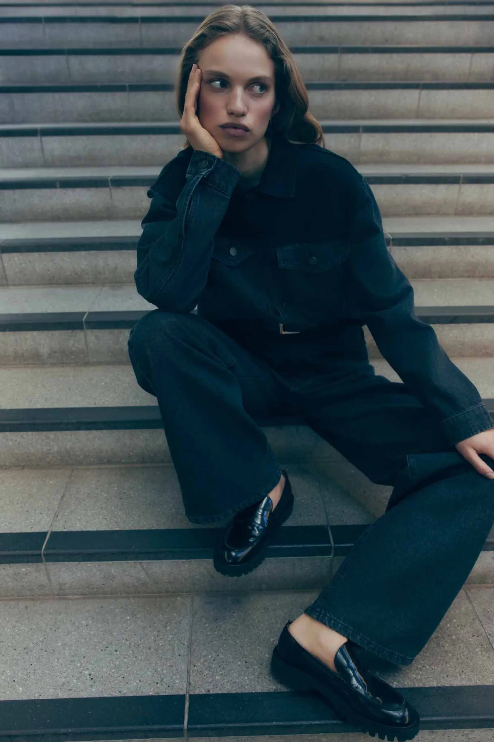 A young woman with wavy brown hair sits on concrete stairs in a dark denim outfit and black loafers, resting her face in her hand.