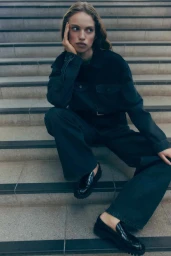 Model seated on concrete stairs wearing a dark denim jacket and matching trousers with black loafers