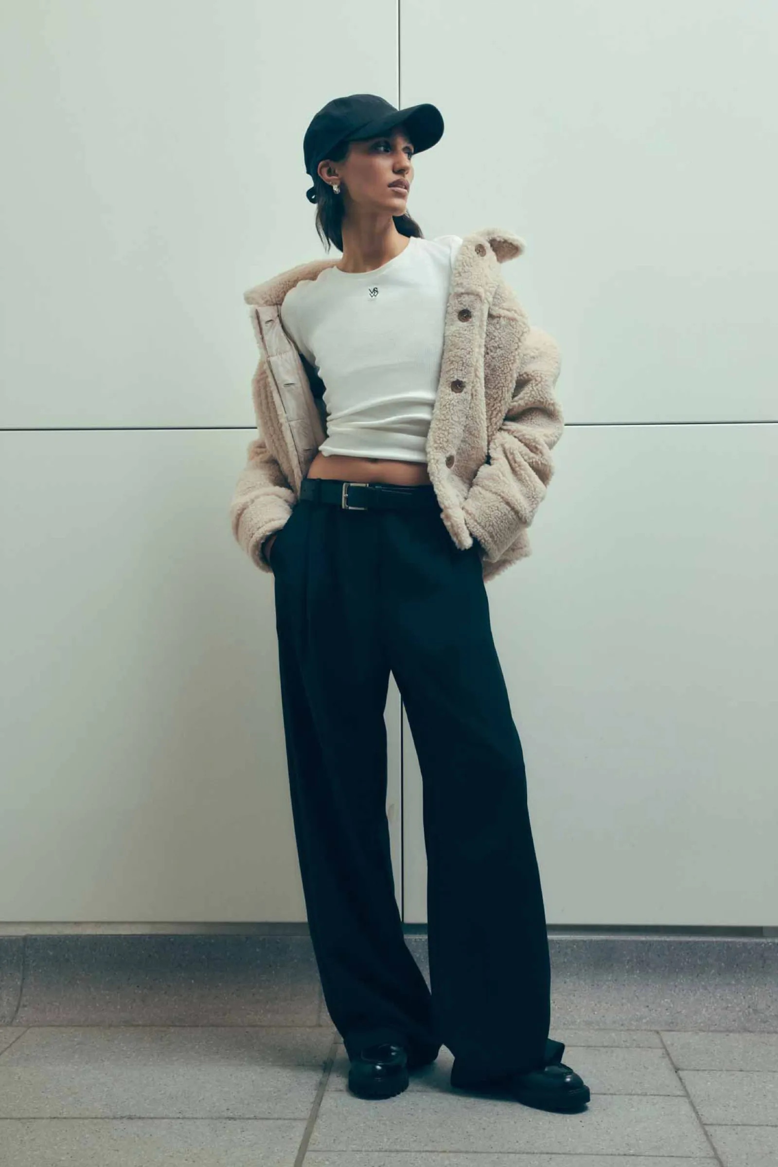 A young woman in a cream-colored jacket, white shirt, dark trousers, and cap poses in front of a white geometric wall indoors.